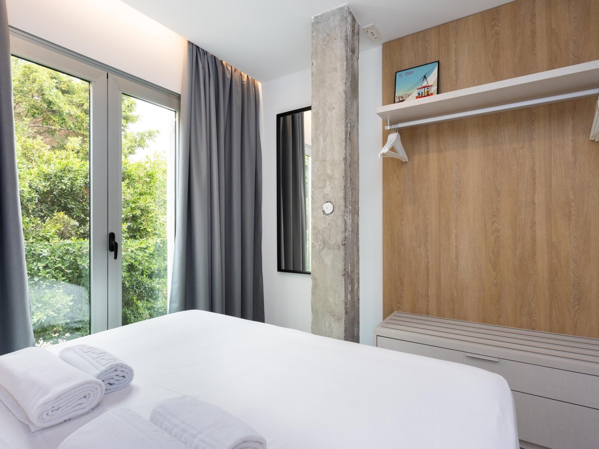 Ein modernes Hotelzimmer mit einem großen Bett, einer Wandpaneele aus Holz, einem Fenster mit Vorhängen und einem kleinen Regal mit einem Bild.