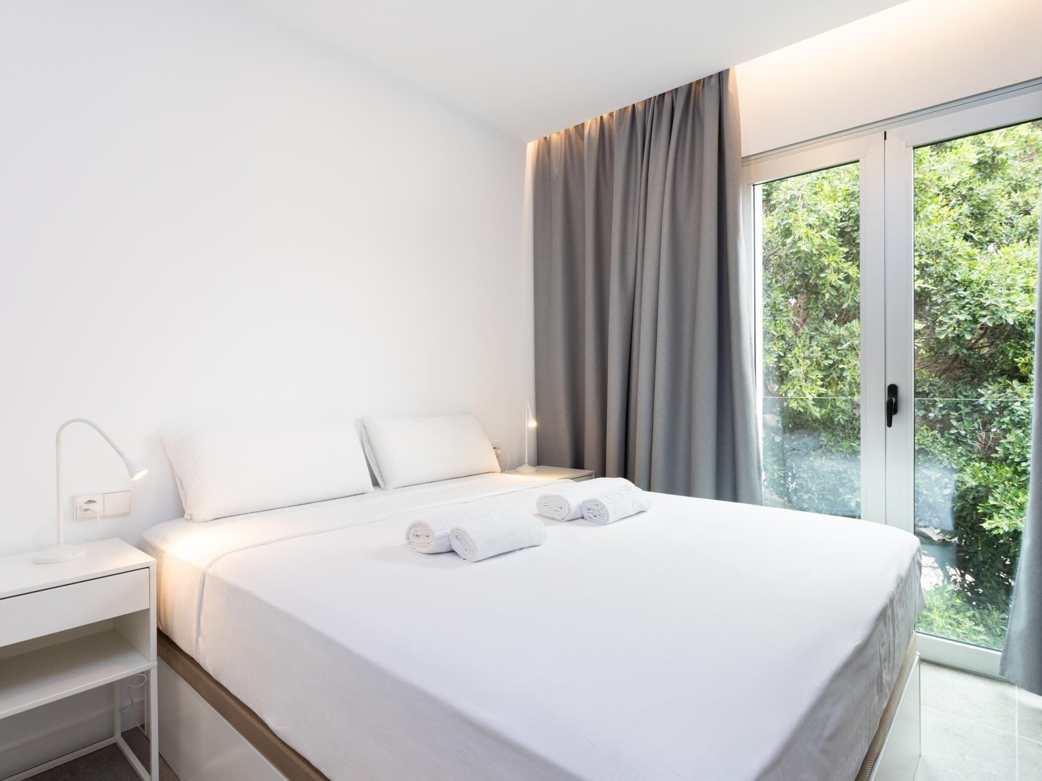 A modern hotel room with a neatly made bed, a bedside table with a lamp, and a view of greenery through the window.