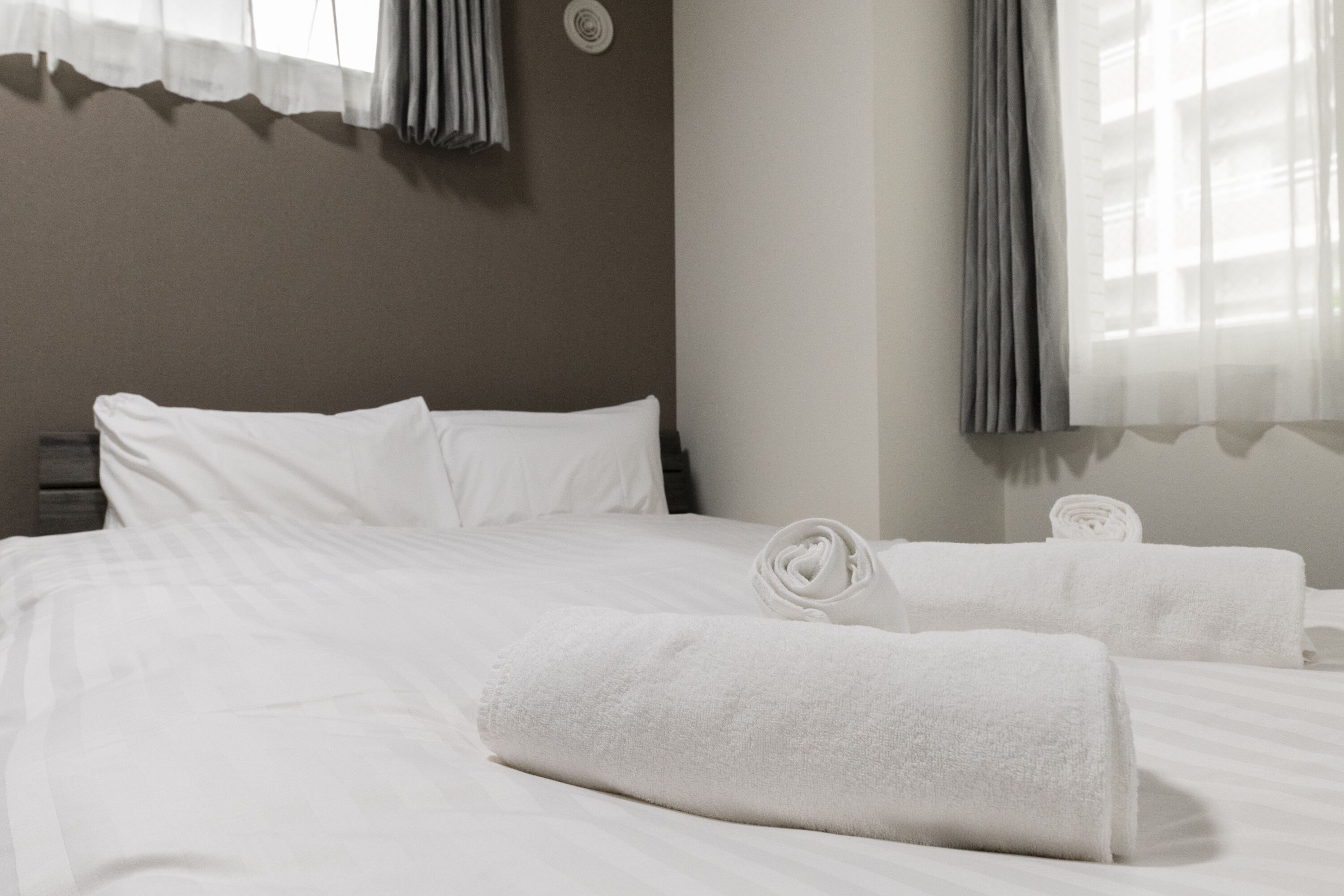 A hotel room with a neatly made bed, two rolled towels, and a window with curtains.