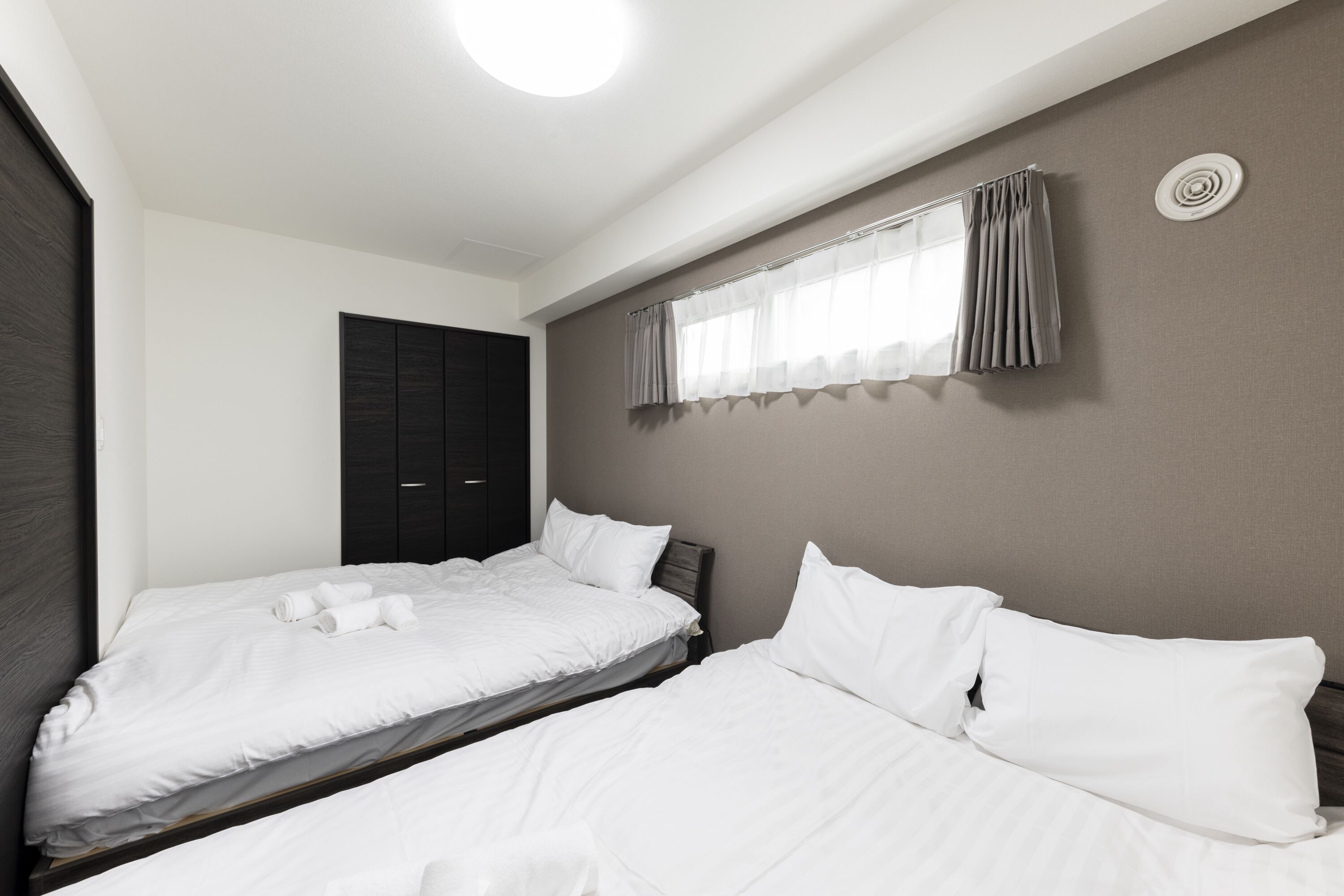 A hotel room with two single beds, a dark wardrobe, a window with curtains, and a ceiling light.