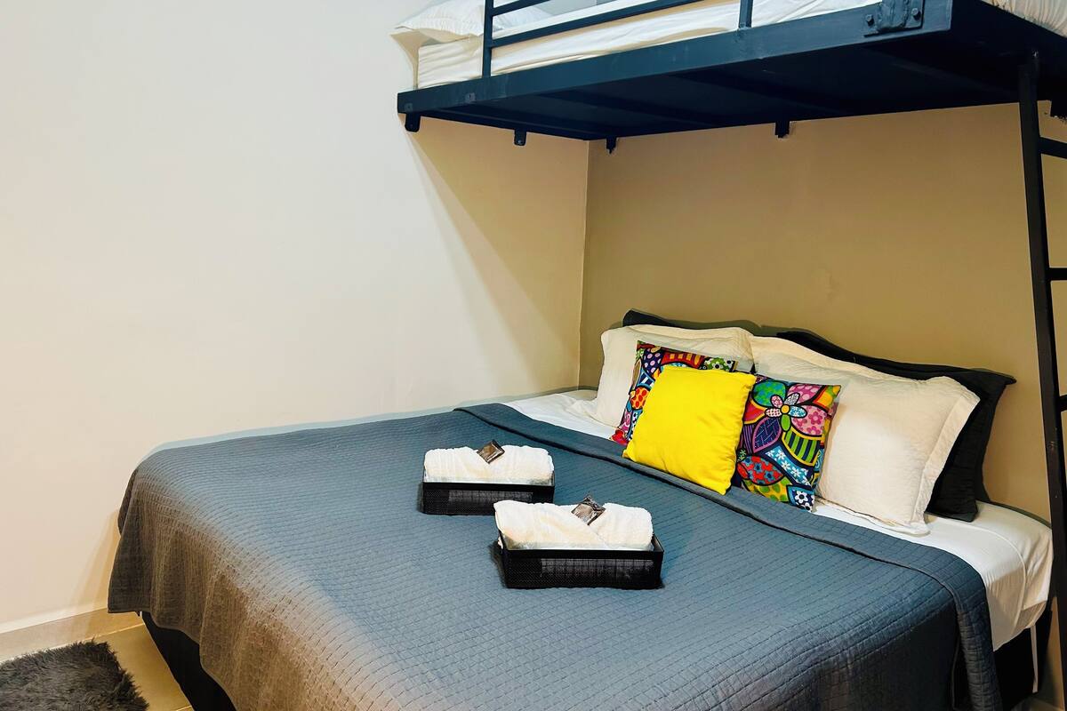 Uma cama beliche com estrutura preta, almofadas brancas e uma almofada colorida. Há duas bandejas pretas com toalhas brancas na cama.