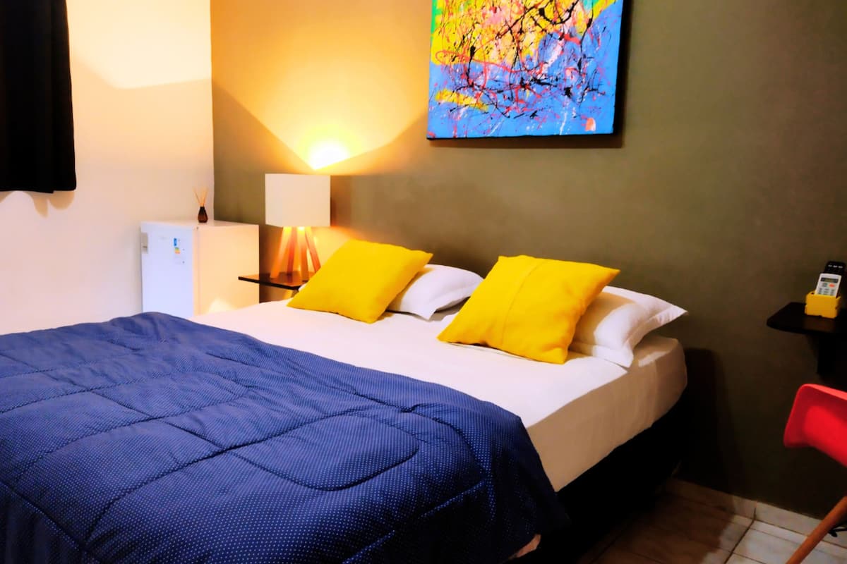 Quarto de hotel com cama, mesa de cabeceira, luminária, pintura e uma pequena mesa com telefone.