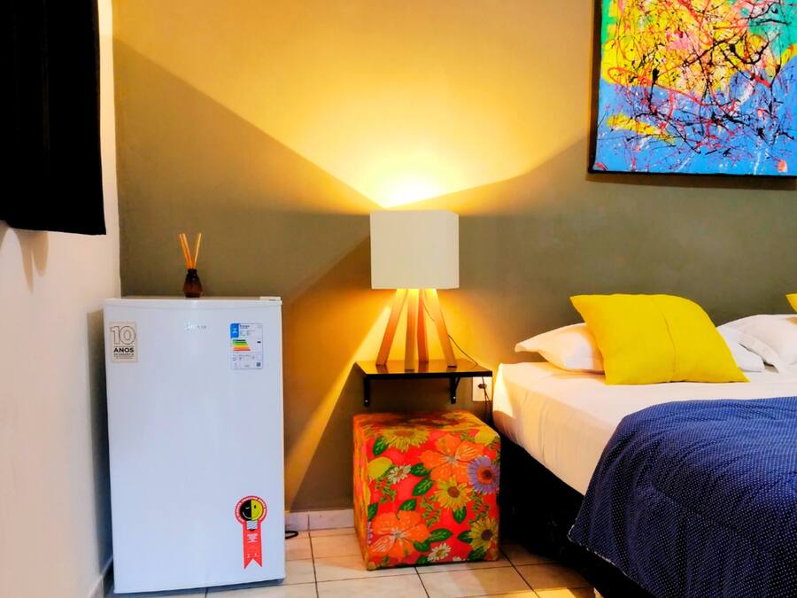 Un dormitorio con cama, mesita de noche, lámpara, refrigerador y un otomano colorido.