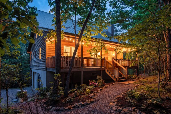 A log cabin with a wooden deck and stairs leading to a balcony, surrounded by trees and a gravel path.