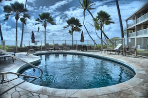 A swimming pool with lounge chairs and umbrellas, surrounded by palm trees and a building with a balcony. - Sea and Sky Beachfront Condo, AC, Ocean Views, Sunrises, Steps to Beach! (Kapaʻa)