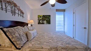 Ein Schlafzimmer mit einem Bett, einem Deckenventilator, einer Wandlampe, einem Gemälde und einem Fenster mit Jalousien.