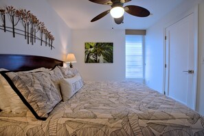 A bedroom with a bed, a ceiling fan, a wall-mounted lamp, a painting, and a window with blinds. - Sea and Sky Beachfront Condo, AC, Ocean Views, Sunrises, Steps to Beach! (Kapaʻa)