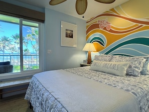 A bedroom with a large bed, a window with a view of palm trees, and a wall with a colorful mural. - Sea and Sky Beachfront Condo, AC, Ocean Views, Sunrises, Steps to Beach! (Kapaʻa)