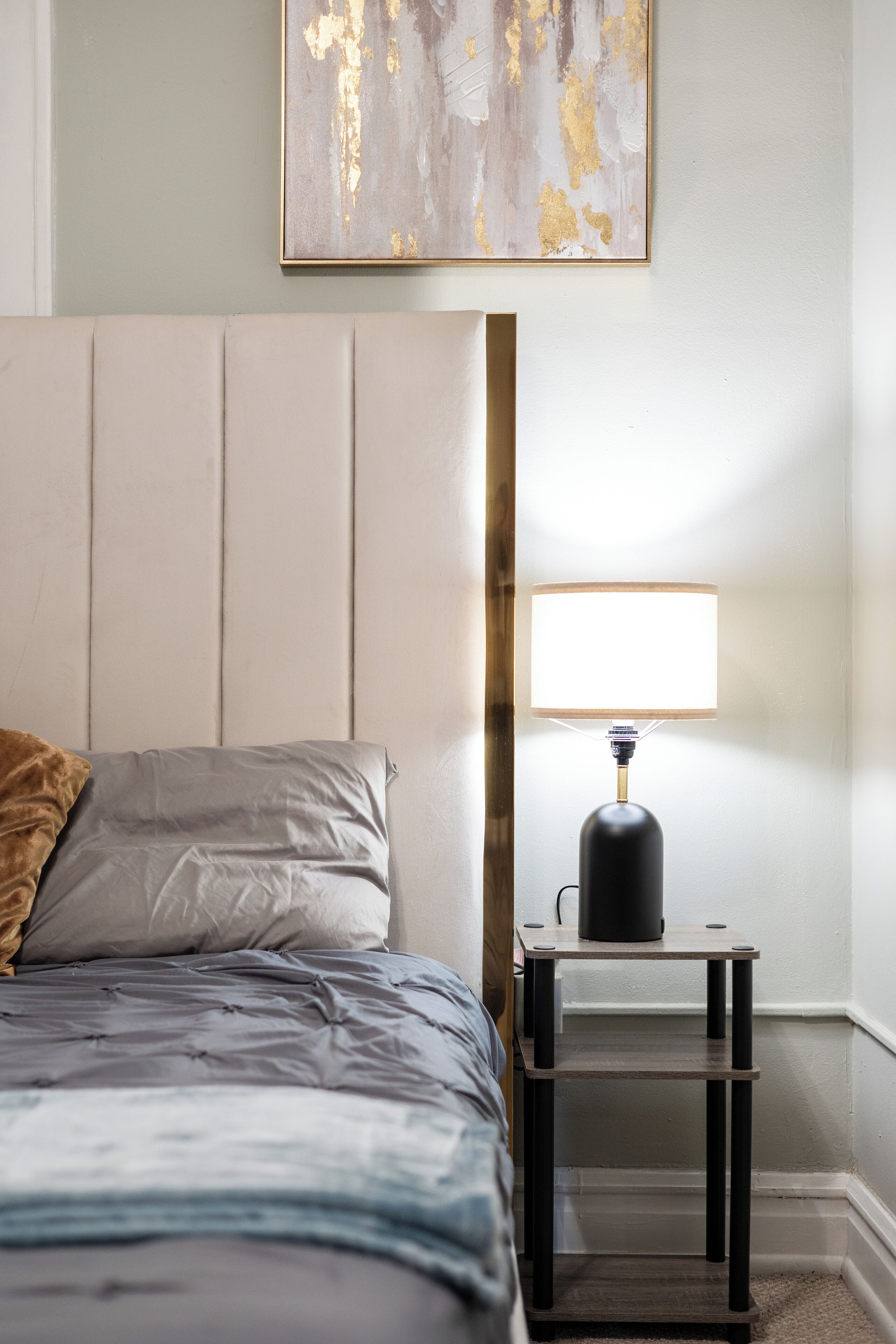 A neatly made bed with a grey and blue duvet, a wooden bedside table with a lamp, and a painting on the wall.