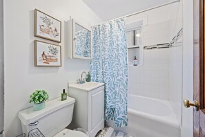 Een badkamer met toilet, wastafel en bad. Er hangen afbeeldingen in lijsten aan de muur en een douchegordijn met een bladmotief.