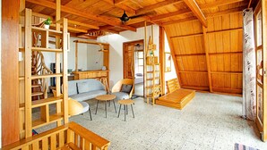 Een ruime kamer met een hoge plafond, houten balken, een bank, twee stoelen, een kleine tafel en een trap naar een hoger niveau.