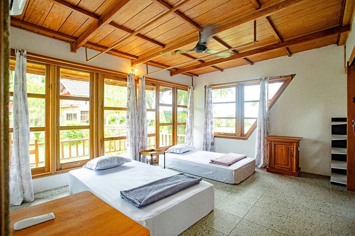 Une chambre avec deux lits, des poutres apparentes en bois au plafond, de grandes fenêtres et un petit meuble en bois.