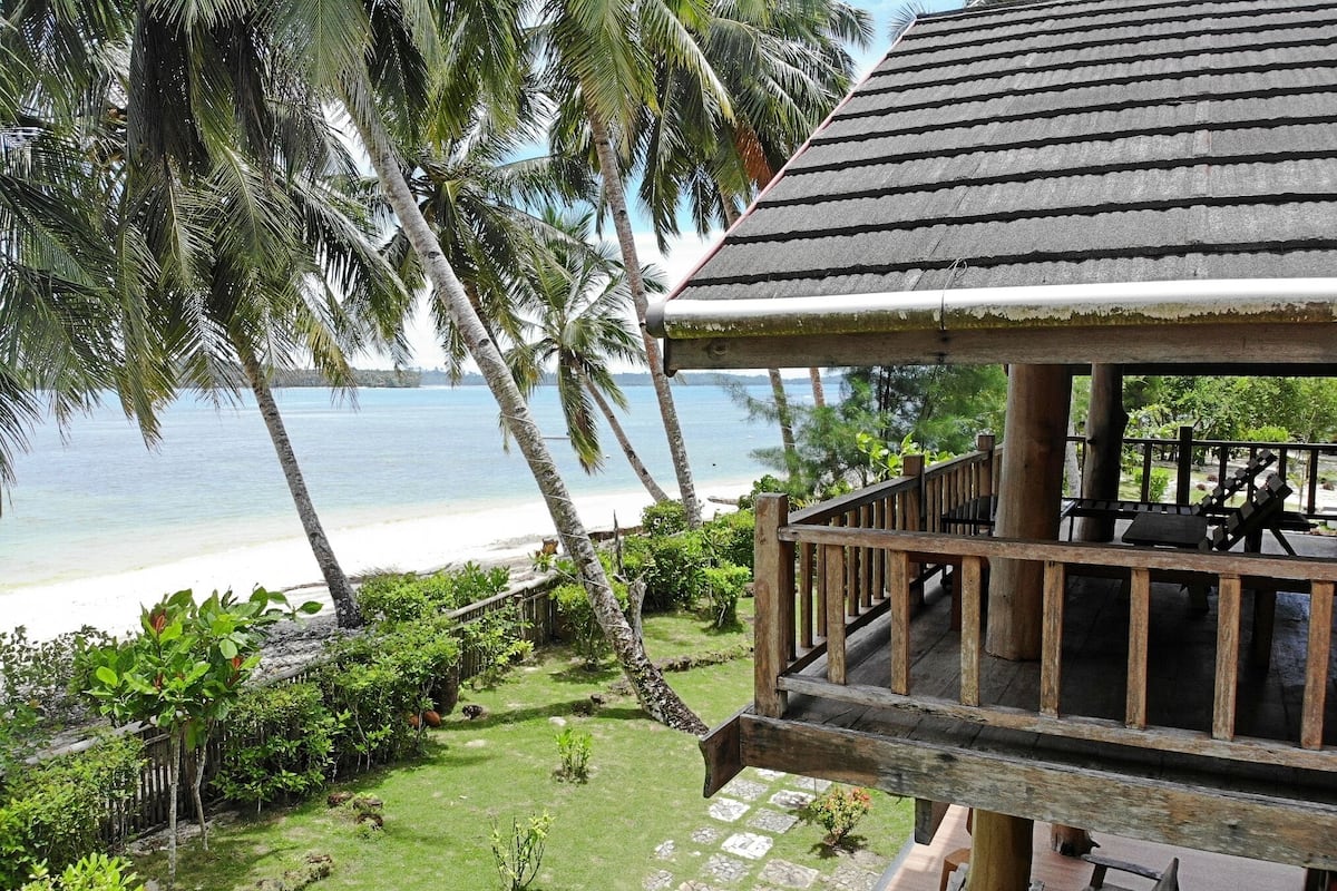 Une terrasse en bois donnant sur une plage bordée de palmiers, avec une vue dégagée sur l’océan.