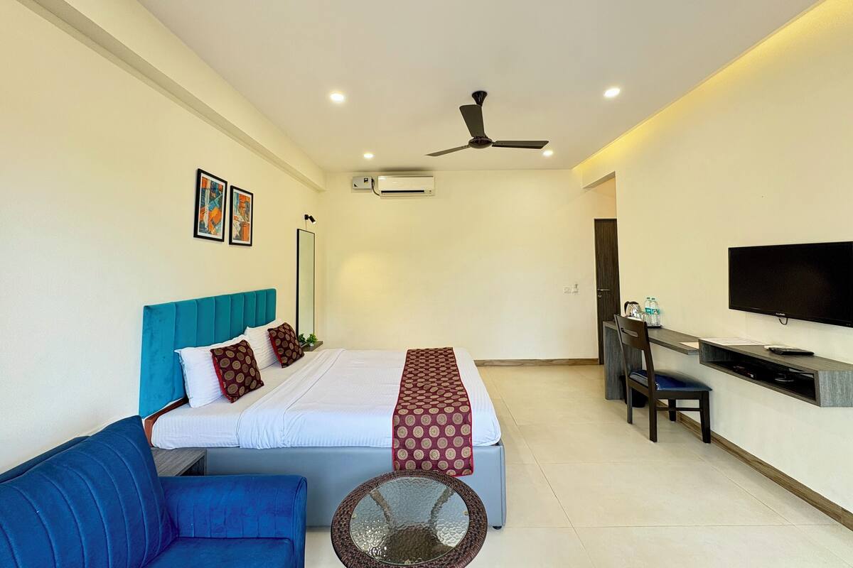 A modern hotel room with a bed, a blue sofa, a desk with a TV, and an air conditioning unit.