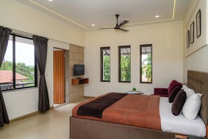 A bedroom with a bed, a ceiling fan, a TV, and windows with curtains. - Girivihar Holiday club - Pure Veg (Mawal)