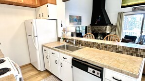 A modern kitchen with white cabinets, a built-in dishwasher, a large island with a sink, and a stone fireplace.