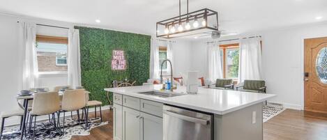 Uma cozinha moderna com uma ilha central, um espaço de refeições com cadeiras, uma parede verde e uma placa com a inscrição 'THIS MUST BE THE PLACE'.