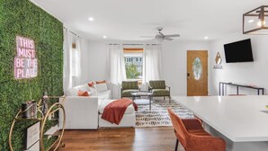 A modern living room with a green wall, white furniture, a coffee table, and a TV mounted on the wall. - Game Room, Balcony, Fire Pit, 5 Bed, 4 Bath, WFH (Charlotte)