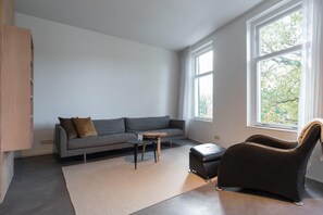 Un salon moderne avec un canapé gris, une petite table en bois, un fauteuil en cuir noir et une grande fenêtre donnant sur des arbres.