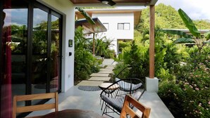 Un patio exterior moderno con una mesa y sillas de madera, rodeado de abundante vegetación y un sendero que lleva a un edificio blanco.