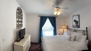 Une chambre à coucher avec un lit, un ventilateur de plafond, un téléviseur posé sur une commode, une lampe et un tableau encadré au mur.