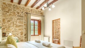 A bedroom with a stone wall, a bed with white linens, a wooden chair, and a window with curtains. - Sa Bisbal by Interhome (Moscari)