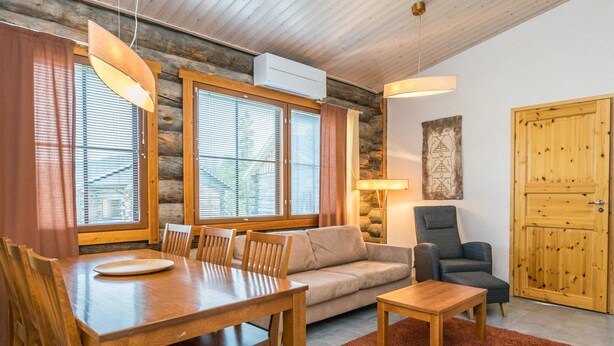 Een gezellige woonkamer met een houten eettafel, bank en stoelen, grote ramen met jaloezieën, een lamp en een houten deur.