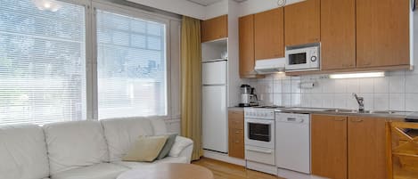 Un espacio habitable compacto con sofá, una pequeña mesa redonda, una kitchenette equipada con microondas, horno y lavaplatos, y una ventana con persianas.