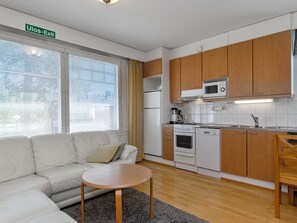 Un espacio habitable compacto con sofá, una pequeña mesa redonda, una kitchenette equipada con microondas, horno y lavaplatos, y una ventana con persianas.