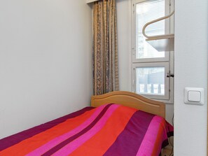 Una cama individual con un edredón colorido a rayas en una habitación con paredes blancas, una ventana con persianas y un cabecero de madera.