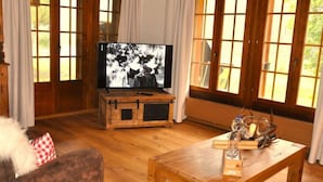 A cozy living room with a wooden TV stand, a flat-screen TV displaying a black-and-white photo, a wooden coffee table with a candle holder, and large windows with curtains.