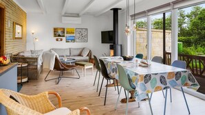 Un moderno comedor con una mesa dispuesta para cuatro personas, rodeada de sillas de mimbre y un sofá. Se ve una chimenea al fondo, y la habitación tiene ventanas amplias con vistas al exterior.