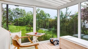 Una habitación iluminada por el sol, con una terraza de madera, una mesa, sillas y un amplio ventanal con vistas a un jardín exuberante.