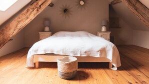 Una camera da letto con soffitto spiovente, travi in legno, un letto con biancheria bianca, due comodini con lampade e un cesto di vimini sul pavimento.