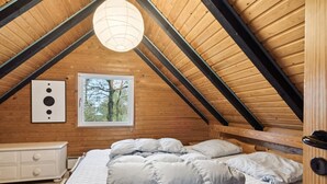 Camera da letto con soffitto spiovente in legno, un letto con molti cuscini, una cassettiera bianca e una finestra vista alberi.