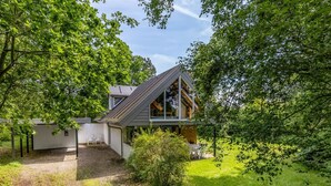 Una casa moderna con tetto spiovente, circondata da vegetazione lussureggiante e un vialetto sterrato.