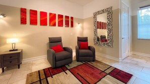 Una habitación con dos sillones grises con cojines rojos, una alfombra estampada, un espejo con marco ornamentado y una pared con arte abstracto rojo.