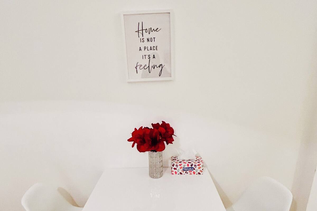 A white dining table with a framed quote on the wall, a vase with red flowers, and a tissue box.