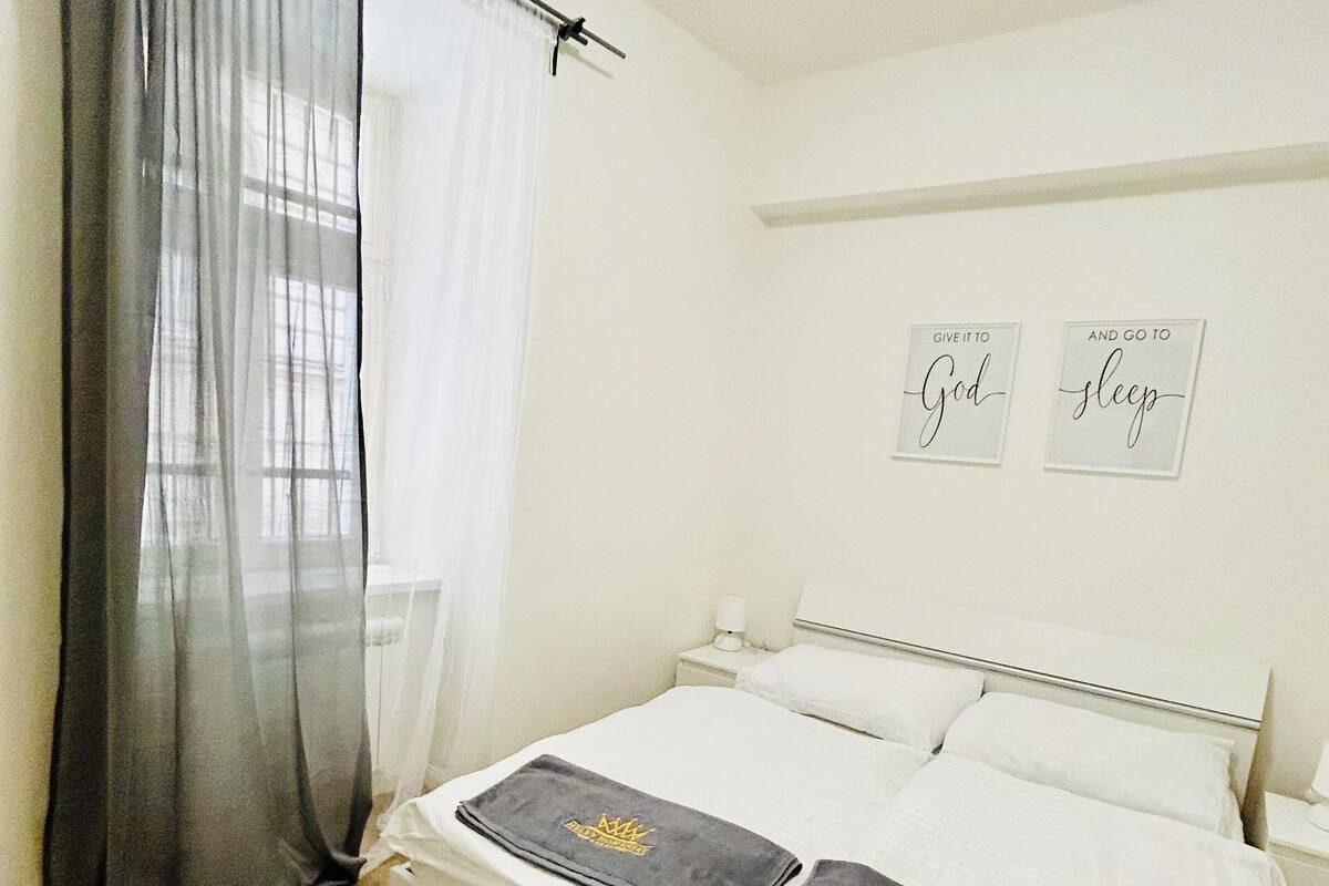 A neatly made bed with white linens and two towels with a crown emblem, a white nightstand with a lamp, a white wall with two motivational signs, and a sheer grey curtain partially drawn.
