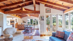 Un vaste salon doté d’un haut plafond en bois, un coin repas avec une grande table et des chaises, et un espace salon confortable avec un canapé bleu et des coussins à motifs.