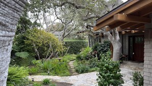 Un chemin de pierre mène à une pergola en bois qui surplombe une terrasse en pierre, le tout étant entouré d’une végétation luxuriante et d’arbres.