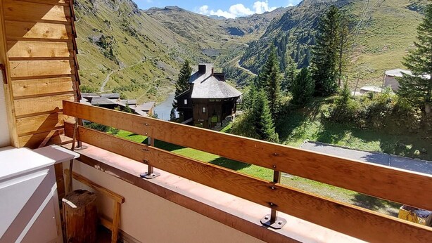 A wooden balcony with a view of a mountain landscape, including a house and a lake.