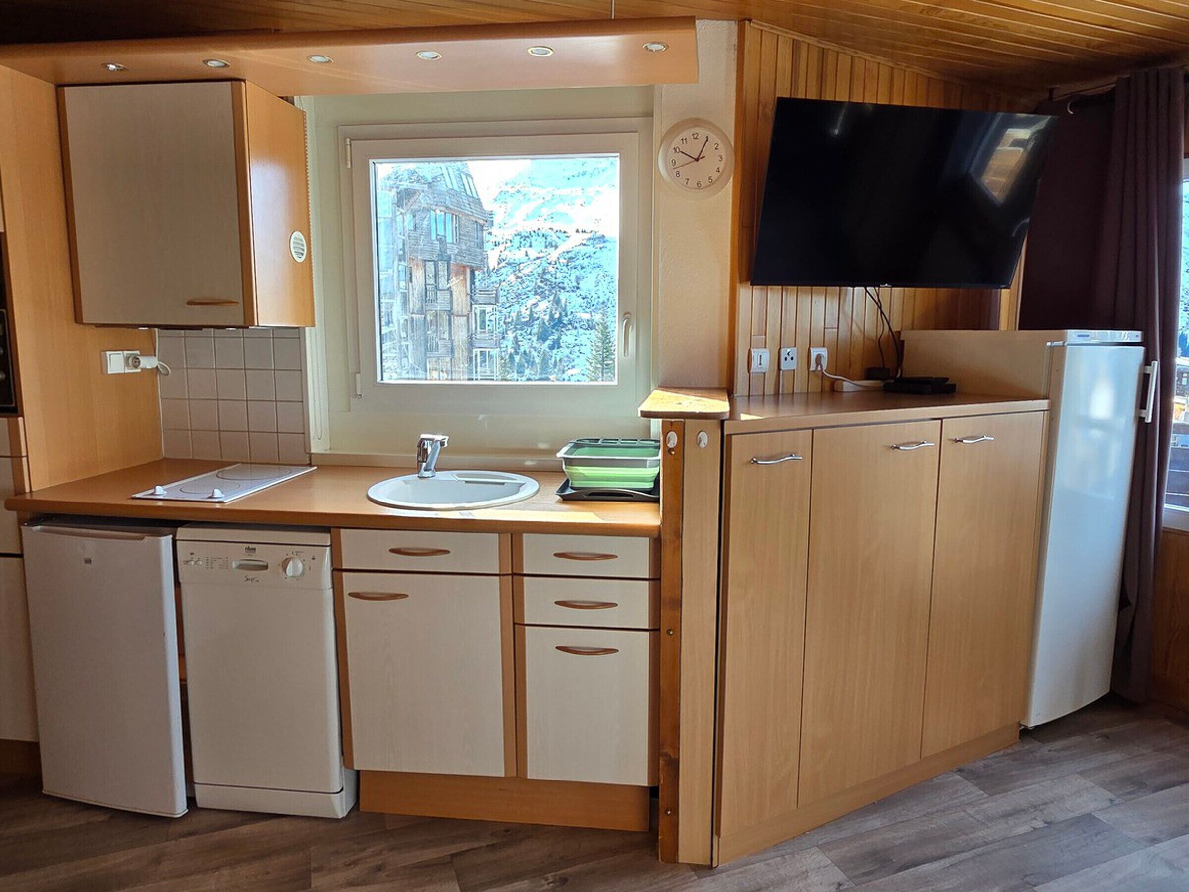 A compact kitchen with wooden cabinets, a sink, a dishwasher, a window with a view, a clock, and a TV mounted on the wall.