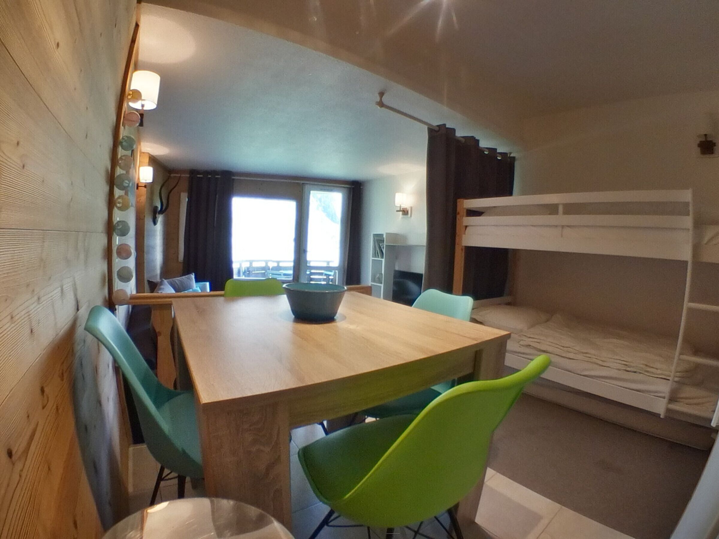 Une petite pièce avec une table en bois, des chaises vertes, un lit superposé et une vue par la fenêtre.