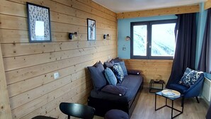 A cozy cabin interior with a wooden wall, a sofa with cushions, a chair, a small table, and a window with curtains.