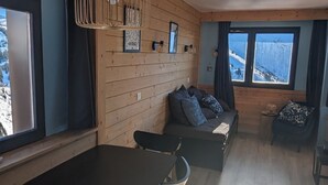 A cozy cabin interior with a wooden wall, a sofa with cushions, a dining table, and a window with a view of a snowy landscape.