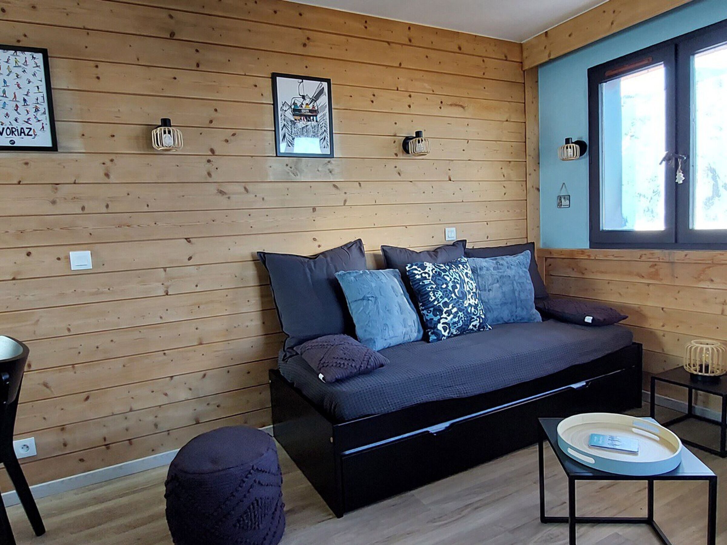 A cozy room with a wooden wall, a sofa with cushions, a small table with a tray, and a window with blinds.