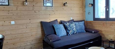 A cozy room with a wooden wall, a sofa with cushions, a small table with a tray, and a window with blinds.