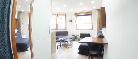 A compact living space with a mirrored wardrobe, a dining table, a sofa, and a small kitchen area.