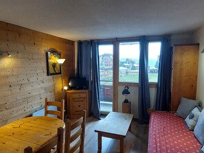 Um quarto aconchegante com paredes de madeira, uma mesa de jantar, uma cama com roupa de cama estampada, uma TV e vista para edifícios pela janela.